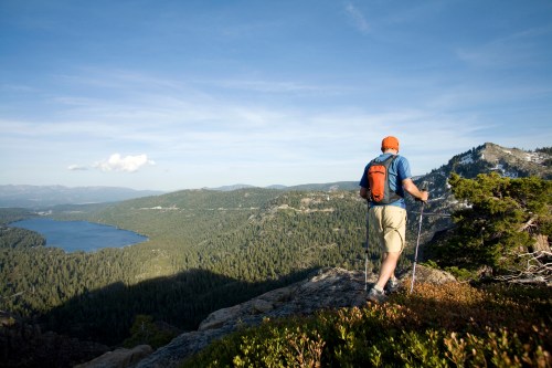A hiker with an orange backpack stands on a rocky outcrop, overlooking a lush valley and a lake in the distance.