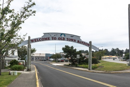 A welcoming sign arching over a road reads "Welcome to Old Town Bandon," with shops and trees in the background.