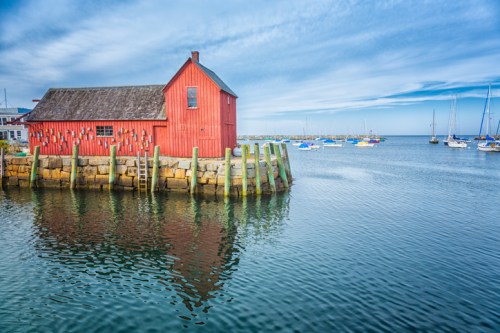 A red wooden building on a pier reflects in calm water, with sailboats in the background under a blue sky.
