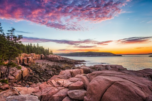 Sunset over rocky coastline with vibrant clouds and trees, reflecting on calm waters in a serene landscape.