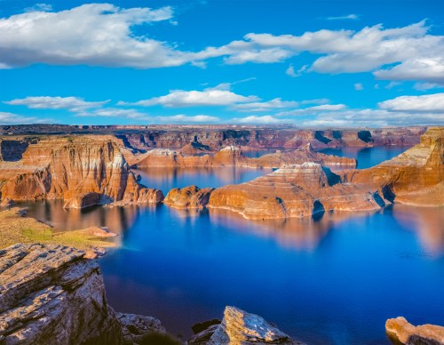 A stunning landscape of red rock formations and blue water under a bright sky with fluffy clouds.