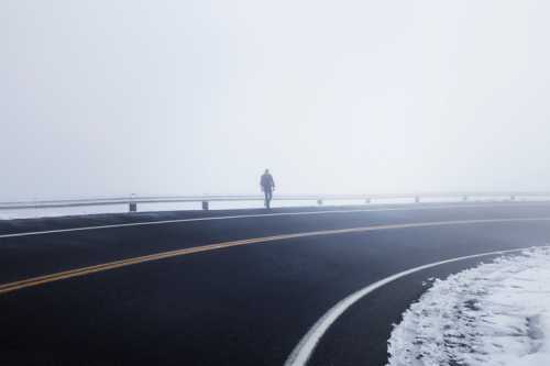 A lone figure walks along a foggy, winding road with snow on the edges, creating a mysterious atmosphere.