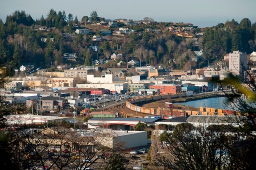 Aerial view of a coastal town with buildings, a train track, and a waterfront, surrounded by hills and trees.