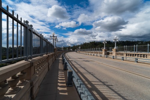 A curved bridge with decorative railings under a cloudy sky, surrounded by greenery.