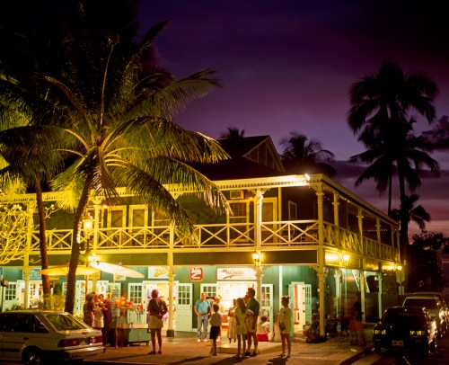 A vibrant evening scene of a tropical building with palm trees, people socializing, and warm lights illuminating the area.