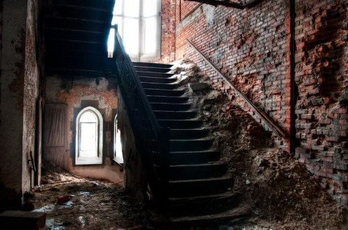 Abandoned interior with a worn staircase, exposed brick walls, and debris scattered on the floor, illuminated by natural light.