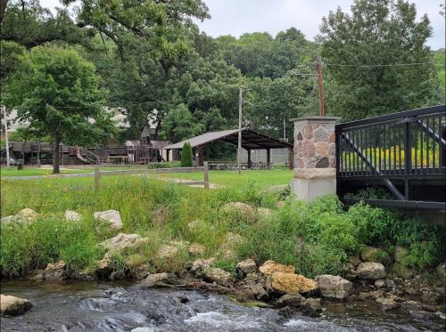 A peaceful scene featuring a stream, grassy area, and a pavilion surrounded by trees and a bridge.