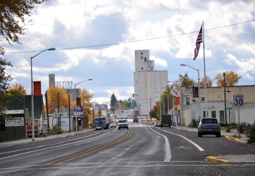 A quiet street in a small town, featuring shops, a grain elevator, and an American flag, under a cloudy sky.