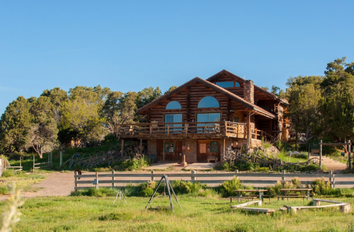 A large log cabin surrounded by greenery, featuring multiple balconies and large windows under a clear blue sky.