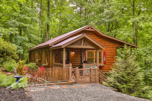 A cozy log cabin surrounded by lush greenery, featuring a front porch and a fenced yard.