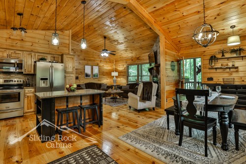 Cozy cabin interior with wooden walls, a kitchen, dining area, and living space featuring large windows and rustic decor.