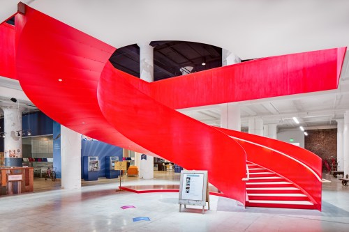 A vibrant red spiral staircase in a modern space, featuring white steps and surrounded by industrial-style architecture.