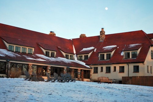 A snow-covered landscape features a large, rustic house with a red roof and a clear sky with a visible moon.