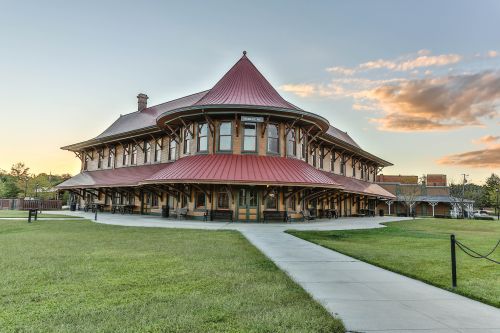 Historic train station with a distinctive red roof, surrounded by green grass and a clear sky at sunset.