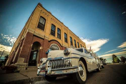 A vintage white car parked in front of a historic brick building under a blue sky with scattered clouds.