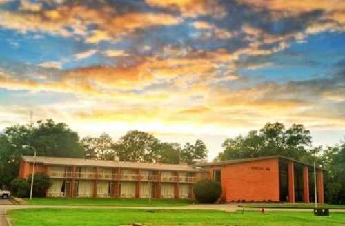 A brick building with multiple rooms, surrounded by green grass and trees, under a colorful sunset sky.