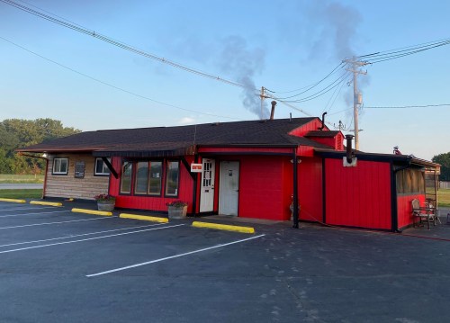 A small, red and wooden building with smoke rising from the chimney, surrounded by a parking lot and greenery.