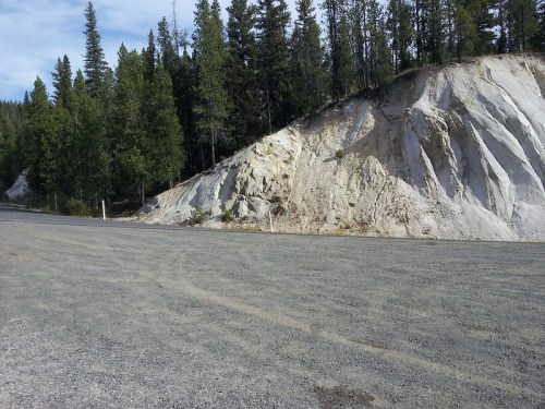A gravel road beside a steep, rocky hillside with trees in the background under a clear blue sky.