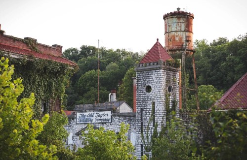 Abandoned distillery with ivy-covered stone walls and a rusty water tower, surrounded by lush greenery.