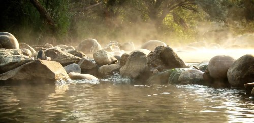 A serene river scene with smooth stones, mist rising from the water, and lush greenery in the background.