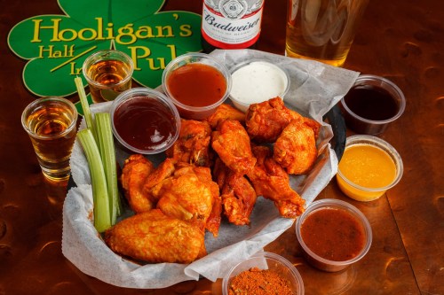 A platter of chicken wings with various sauces, celery sticks, and drinks on a table at Hooligan's Pub.