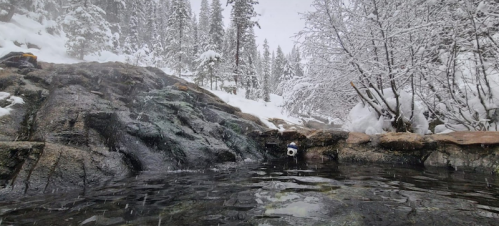 A serene hot spring surrounded by snow-covered rocks and trees, with gentle snowfall in a winter landscape.