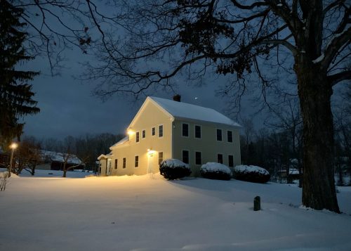A snow-covered house at dusk, illuminated by warm lights, surrounded by trees and a quiet winter landscape.