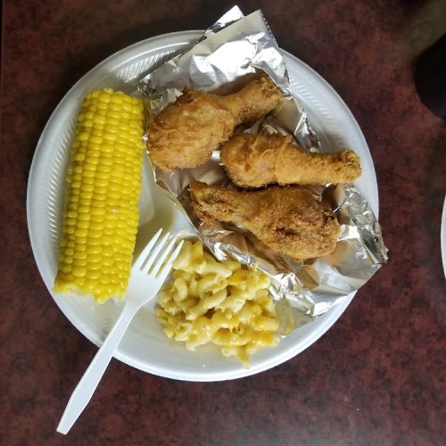 A plate with three fried chicken drumsticks, corn on the cob, and macaroni and cheese, served with a plastic fork.
