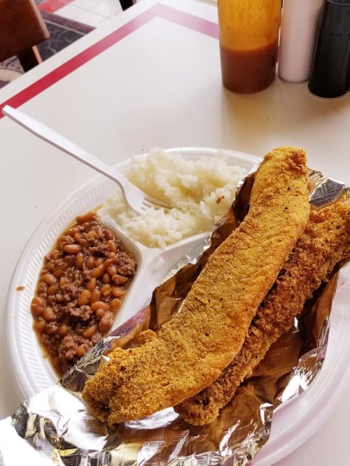 A plate with two fried fish fillets, rice, and baked beans, served with a fork on a white tray.