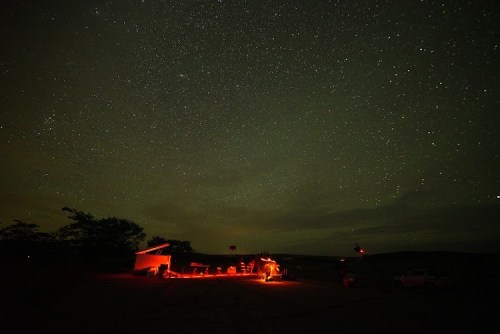 A dark sky filled with stars above a dimly lit building and silhouettes of people gathered outside.
