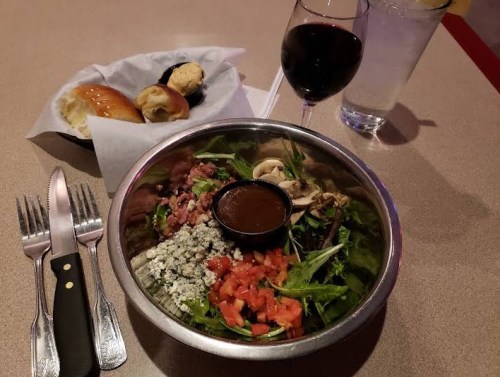 A bowl of salad with toppings, a glass of red wine, water, and rolls on a table with utensils.