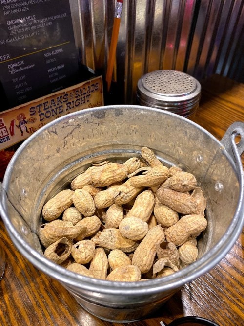 A metal bucket filled with unshelled peanuts on a wooden table.
