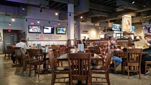 A casual restaurant interior with wooden tables, chairs, and TVs on the walls, featuring diners enjoying their meals.