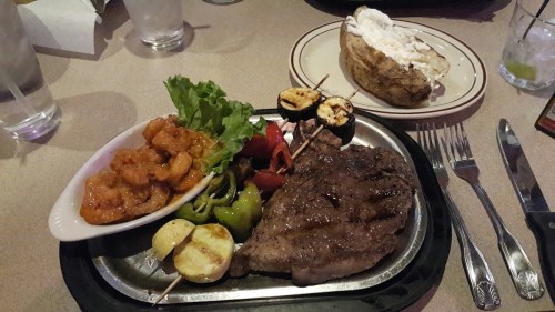A plate with grilled steak, shrimp, vegetables, and a baked potato on the side, set on a table with drinks.