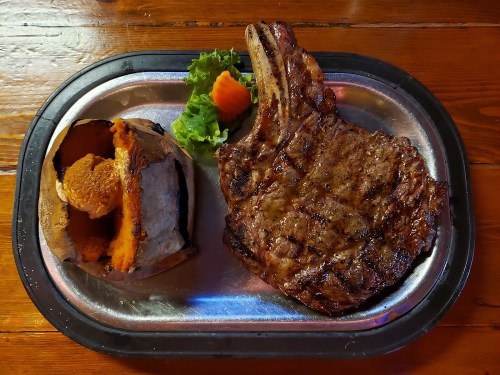A grilled ribeye steak on a metal platter, accompanied by a baked sweet potato and a small salad.