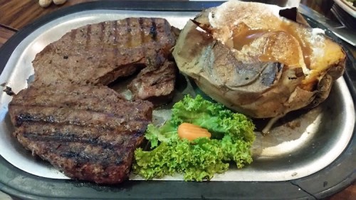 A sizzling platter with grilled steak, a baked potato, and a side of lettuce and carrot.