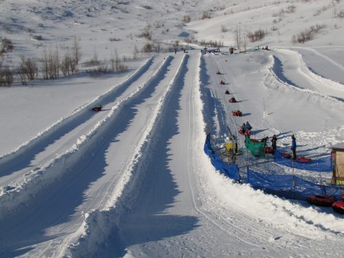 Snowy landscape with sledding tracks, people enjoying sledding, and a blue safety barrier in the background.