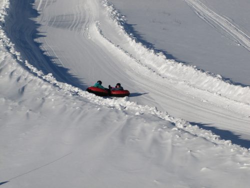 Two people in colorful snow tubes slide down a snowy hill, surrounded by white snow and winding tracks.