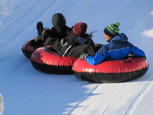 Three people in winter gear are sitting on red snow tubes, sliding down a snowy slope.