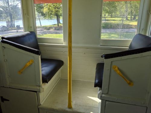Interior of a vintage train car with two black seats, a yellow pole, and windows showing a park view outside.
