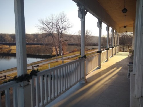 A sunlit porch with white railings overlooks a calm river and bare trees in the distance.
