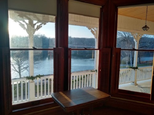 View from a window with wooden frames, overlooking a serene lake and trees in the distance, bathed in soft sunlight.