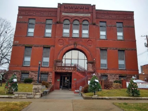 Historic red brick building with an arched entrance, flanked by small evergreen trees, on a cloudy day.