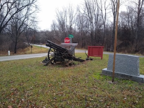 A historic cannon displayed on grass near a stop sign and a stone monument, surrounded by bare trees.