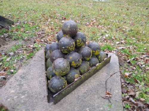 A pyramid of weathered cannonballs resting on a stone base, surrounded by grass and fallen leaves.