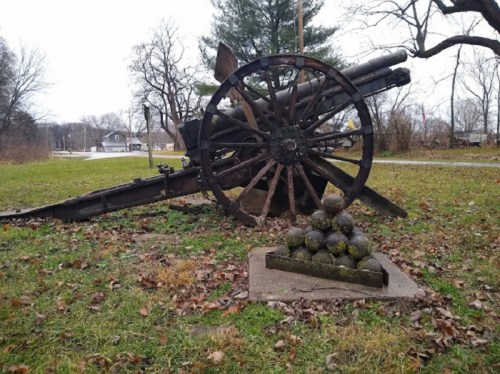 An old cannon on a grassy area, with a stack of cannonballs nearby, surrounded by bare trees and a cloudy sky.