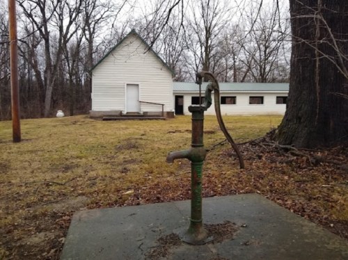An old hand pump in the foreground with a white building and bare trees in the background on a cloudy day.