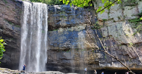 A stunning waterfall cascades down rocky cliffs, surrounded by lush greenery and visitors exploring the area below.