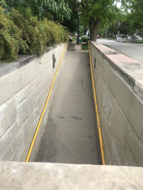 A narrow, paved pathway between stone walls, lined with greenery, leading to a street. A yellow pipe runs along the side.