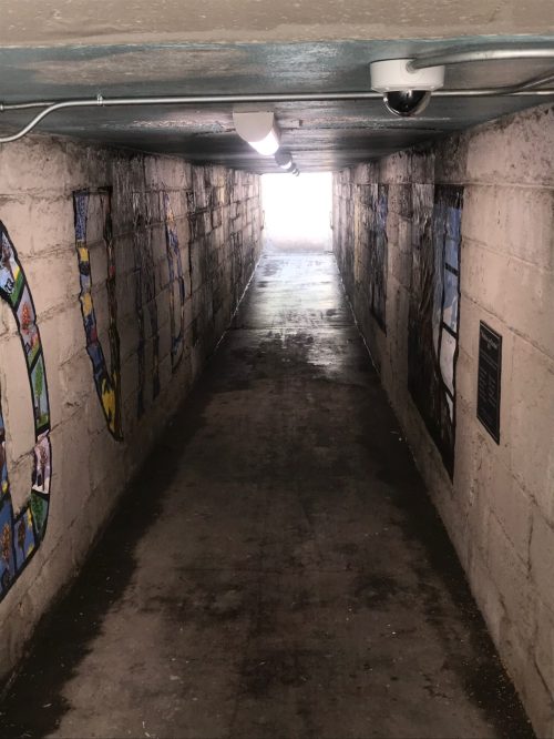 A dimly lit tunnel with murals on the walls, leading to a bright light at the end.